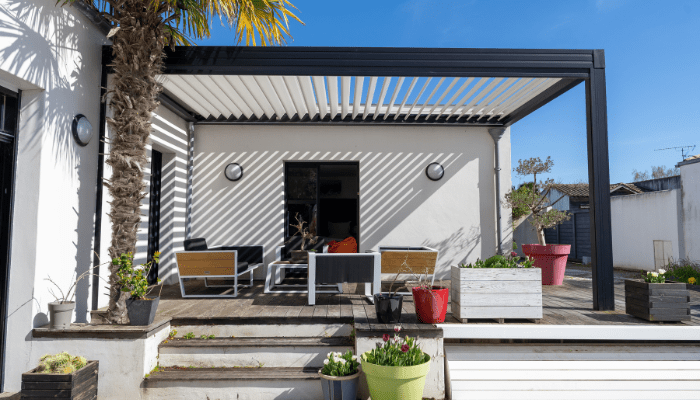 Moderne Terrasse mit Pergola aus Aluminiumlamellen, gemütlicher Sitzgruppe und Blumenkübeln im sonnigen Garten