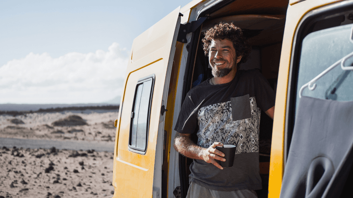 Lächelnder Mann mit Kaffeebecher steht an der offenen Tür eines gelben Campers in einer Wüstenlandschaft